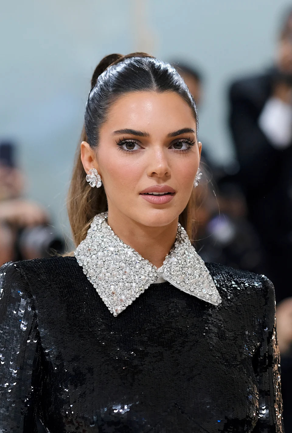 Kendall Jenner in a sequined outfit with a jeweled collar at a formal event, wearing stud earrings, with her hair styled in a sleek ponytail