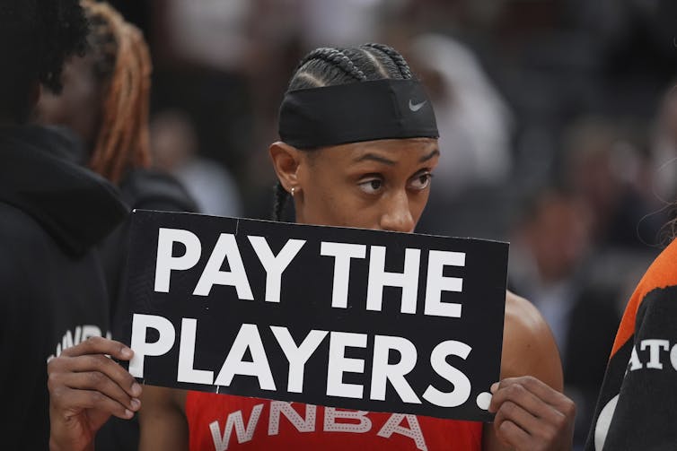 A Black woman with cornrows wearing a black headband and a basketball uniform holds a sign that says 'pay the players'