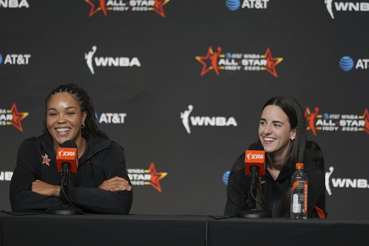 A black woman sits beside a white woman at a conference table in front of a backdrop with the WNBA logo on it