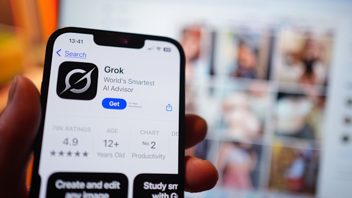 The Grok app page is shown on an iPhone in the App Store, with blurred X (formerly Twitter) search results visible on a laptop screen in the background.
