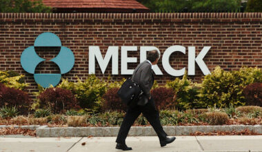 A man carrying a bag walks past a large Merck sign outside a brick building surrounded by shrubs