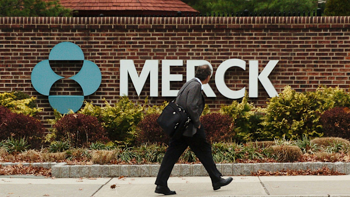A man carrying a bag walks past a large Merck sign outside a brick building surrounded by shrubs