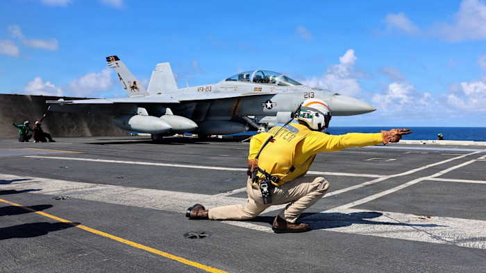 A U.S. Navy F/A-18F Super Hornet prepares to launch from the USS Gerald R. Ford as a crew member in a yellow shirt signals on the flight deck.