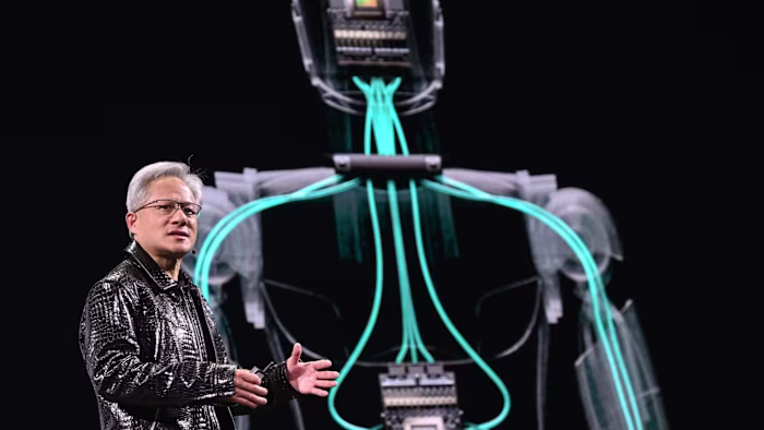 Jensen Huang gestures on stage during his keynote, with a digital illustration of circuitry in a human shape projected behind him.