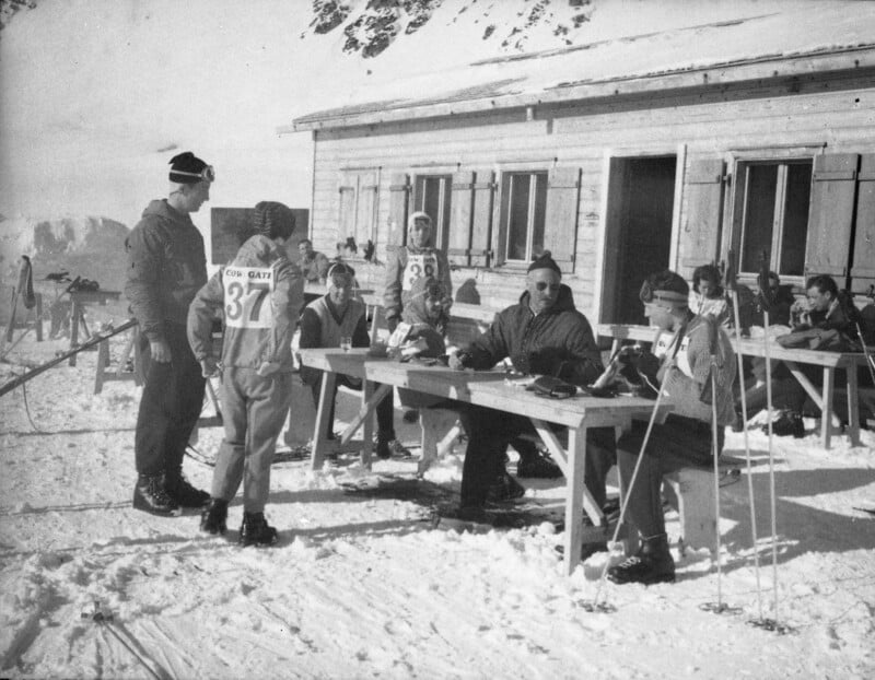 A group of skiers and officials gather outside a wooden cabin in snowy mountains. Some sit at tables with papers, others stand holding ski poles, all wearing winter clothing and numbered bibs. Skis are propped nearby.