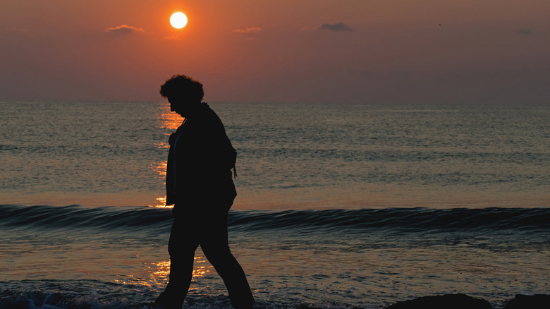 Sad looking older man walking along a waterfront with sunset in background