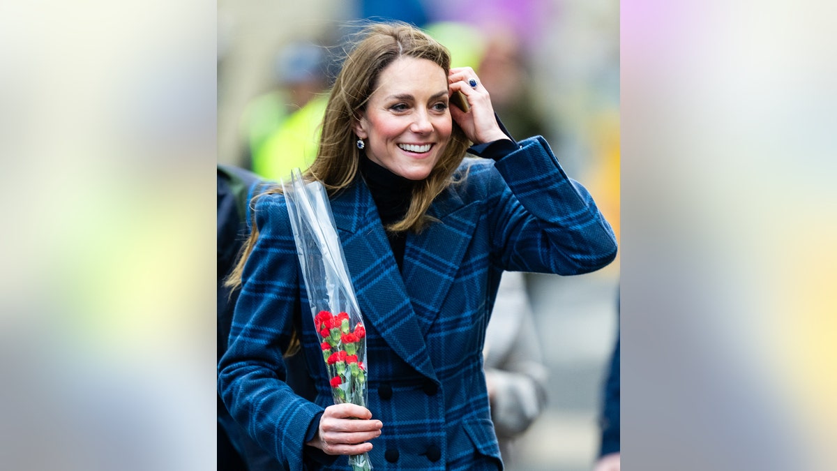 Kate Middleton smiling wearing a blue tartan coat holding a bouquet of flowers.