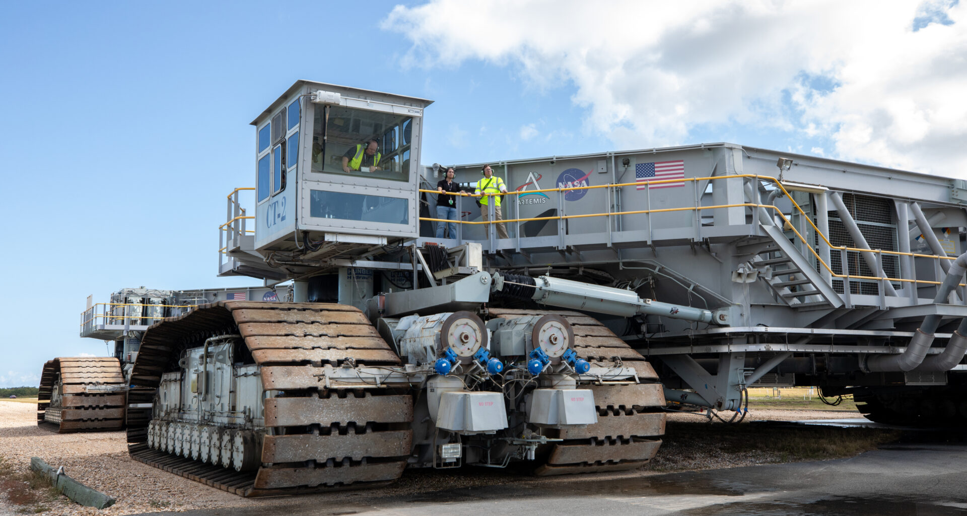 A crawler-transporter, including its massive treads take up the majority of the image. In fact, the entire crawler cannot be seen. There are a few people at the top of the crawler, which allows you to see just how massive the machine is.