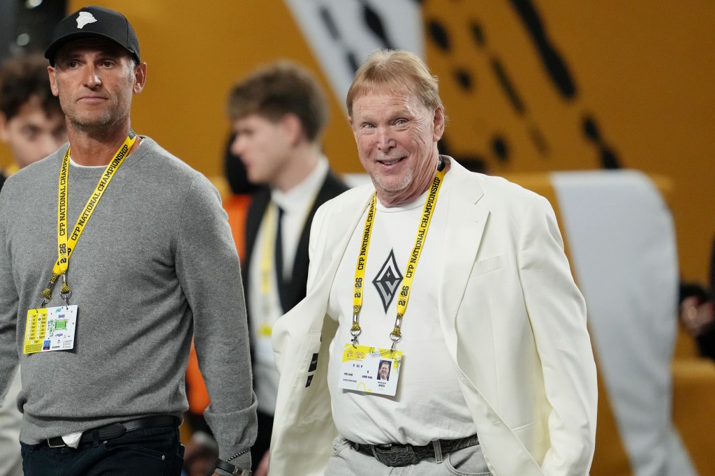 Las Vegas Raiders owner Mark Davis, wearing a white blazer and t-shirt, smiles while walking on the sidelines with another man in a gray sweater and black hat.