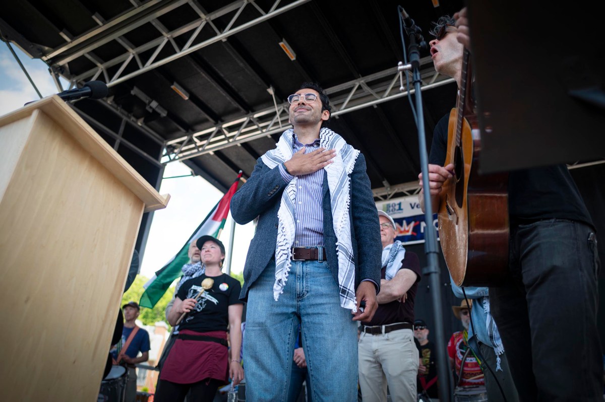 A man with a keffiyeh stands on an outdoor stage with his hand on his chest, surrounded by others; a guitar player and podium are also visible.