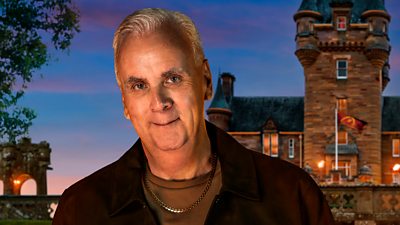 A man in a dark jacket and brown t-shirt smiles to camera, set against the backdrop of a castle by night