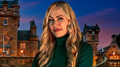 A woman with long blonde hair, wearing a green polo neck top, smiles to camera, set against the backdrop of castle at night