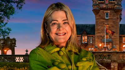 A woman in a green top smiles to camera, standing outside a castle by night
