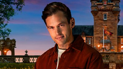 A man in a light blue t-shirt and red shirt jacket smiles to camera, side on, set against the backdrop of a castle by night