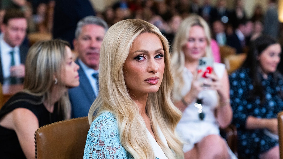 Paris Hilton looking serious as she sits in front of a group of people wearing a light blue lace dress.