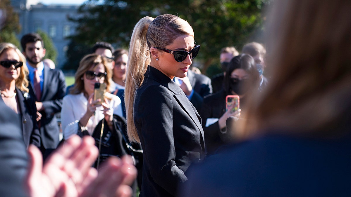 Paris Hilton looking downcast in a black suit with matching sunglasses outdoors.