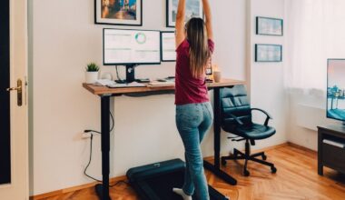 A person in jeans and a dark pink-purple tee using an under-desk treadmill while working on the computer.