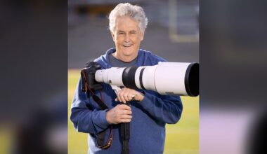 An older man with gray hair, wearing a blue hoodie, smiles while holding a large professional camera with a long telephoto lens on a sports field.