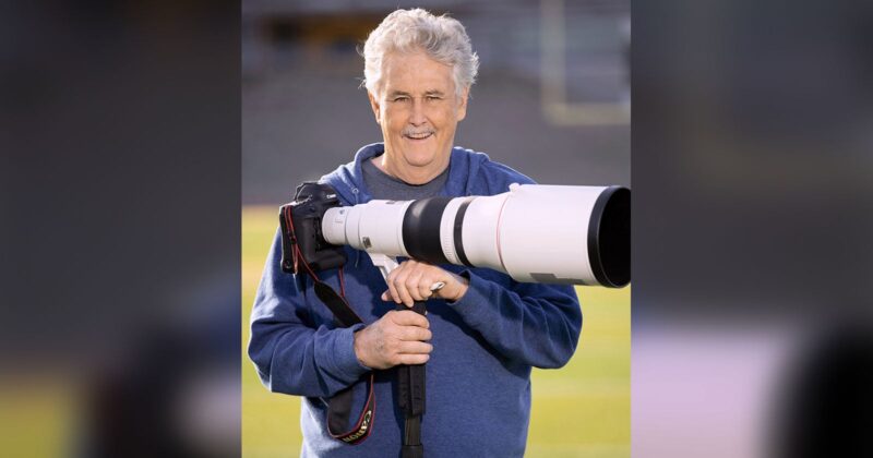 An older man with gray hair, wearing a blue hoodie, smiles while holding a large professional camera with a long telephoto lens on a sports field.
