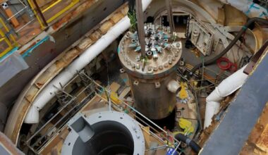 A large cylindrical naval nuclear reactor component is being lifted inside a shipyard work area during maintenance or removal.