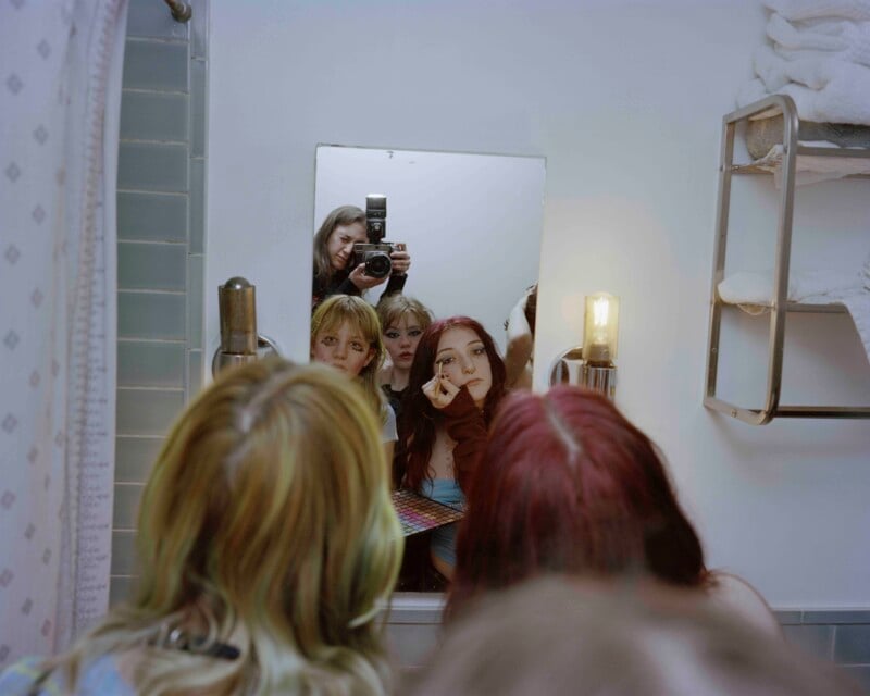 Four young people stand close together in a bathroom, looking into a mirror. One applies makeup, while another takes a photo with a camera. Towels are stacked on a shelf to the right, and the scene is warmly lit.