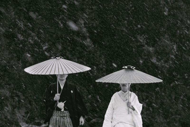 Two people wearing traditional Japanese attire and holding large paper umbrellas walk in falling snow. The background is dark, highlighting the contrast with their clothing and the snowfall.