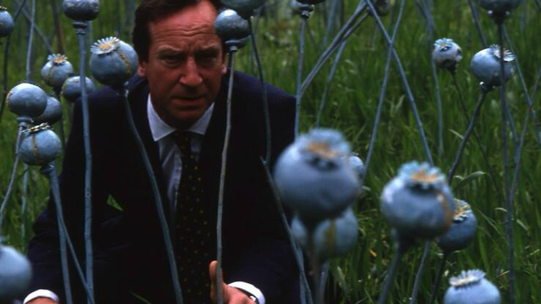 Bill Paterson as Minister Jack Lithgow wearing a suit and tie while kneeling among the blue poppy flowers in 