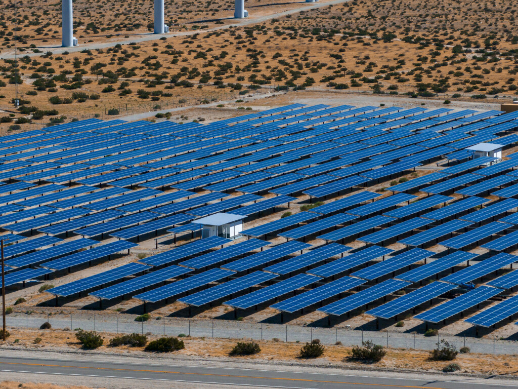 Expansive solar energy farm in Palm Springs, USA, with rows of large blue panels and white wind turbines under a clear sky, amidst a desert landscape.