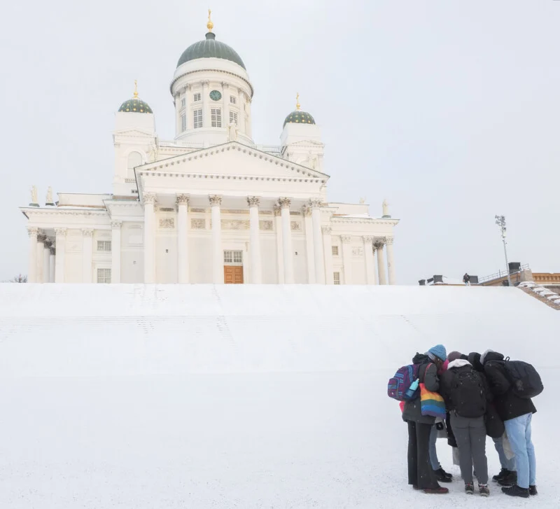 A group of five people with backpacks huddle together in front of the snow-covered Helsinki Cathedral, a large white building with green domes and golden accents.