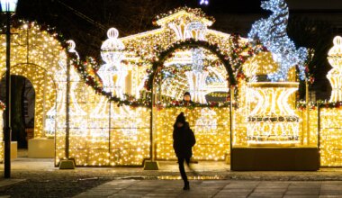 A person in winter clothing walks past festive holiday decorations made of bright yellow and white lights, featuring arches and fountains, on a city street at night. A man looks at her from behind.