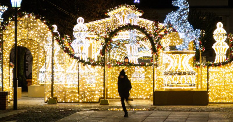 A person in winter clothing walks past festive holiday decorations made of bright yellow and white lights, featuring arches and fountains, on a city street at night. A man looks at her from behind.