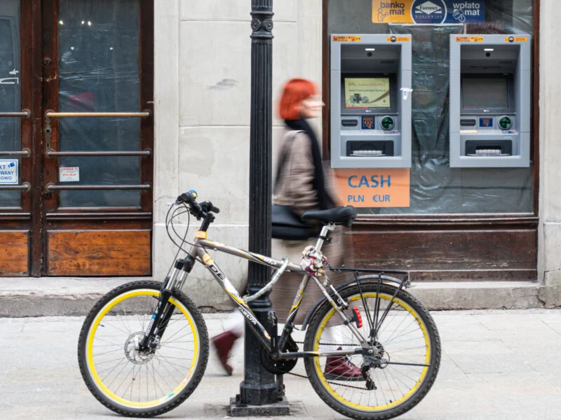 A bicycle is locked to a lamppost on a city sidewalk. A person with red hair walks by, blurred in motion, wearing a mask. Two ATMs are mounted on the wall behind them.