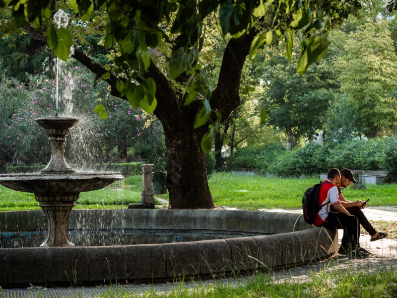 Two people sit on the edge of a circular stone fountain in a lush, green park, shaded by large trees. Sunlight filters through the leaves, and water splashes from the fountain's top tier.