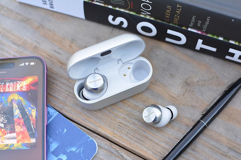 A pair of Technics AZ100 earbuds sit on a wooden desk, with an iPhone and a pair of books next to them. 