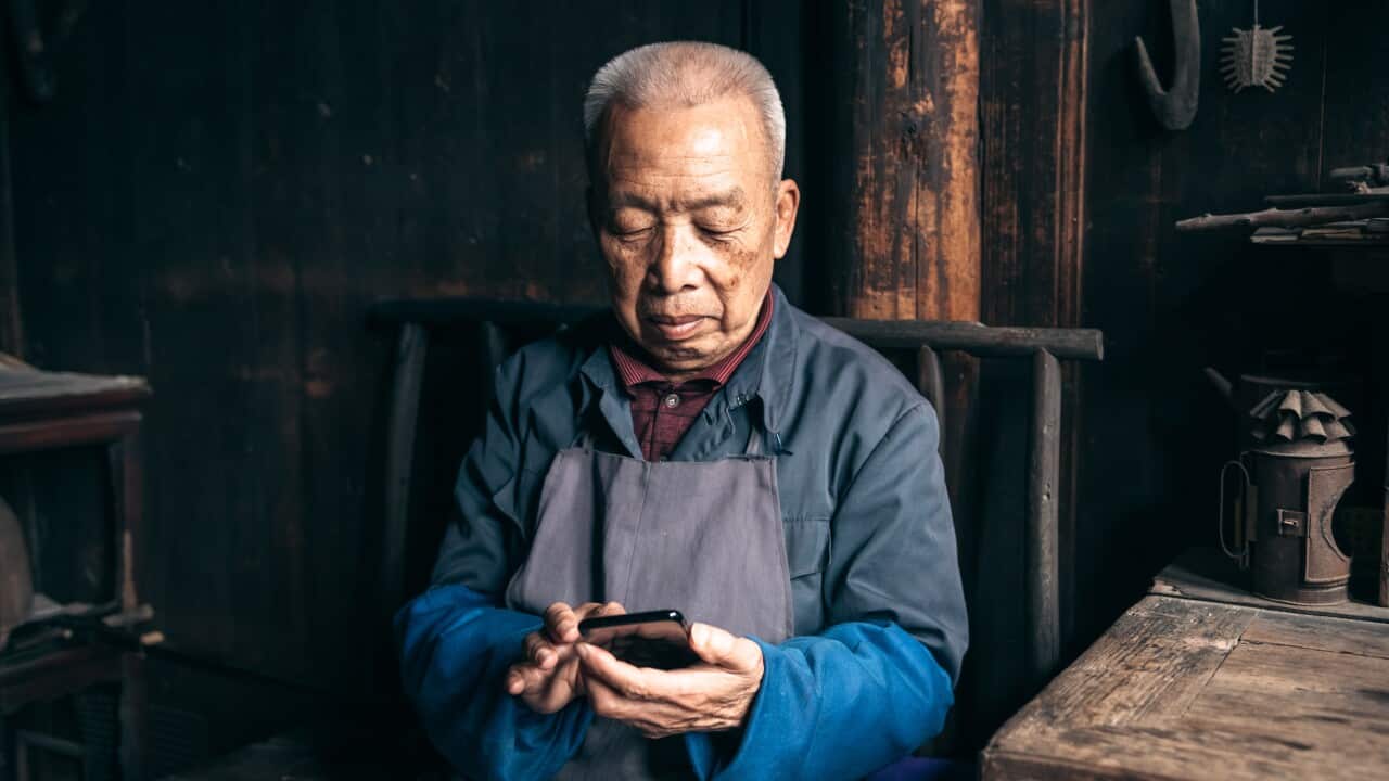 An elderly man in a workshop setting, wearing a blue shirt and gray apron, sits focused on his smartphone against a backdrop of traditional wooden architecture and tools.