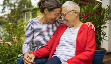 A grown child with a parent are seated, smiling and holding hands.
