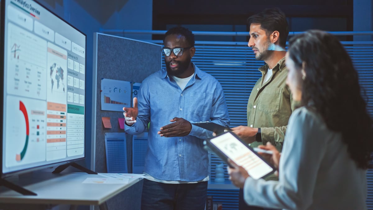 Three people standing in front of a monitor and discussing a chart on the screen.