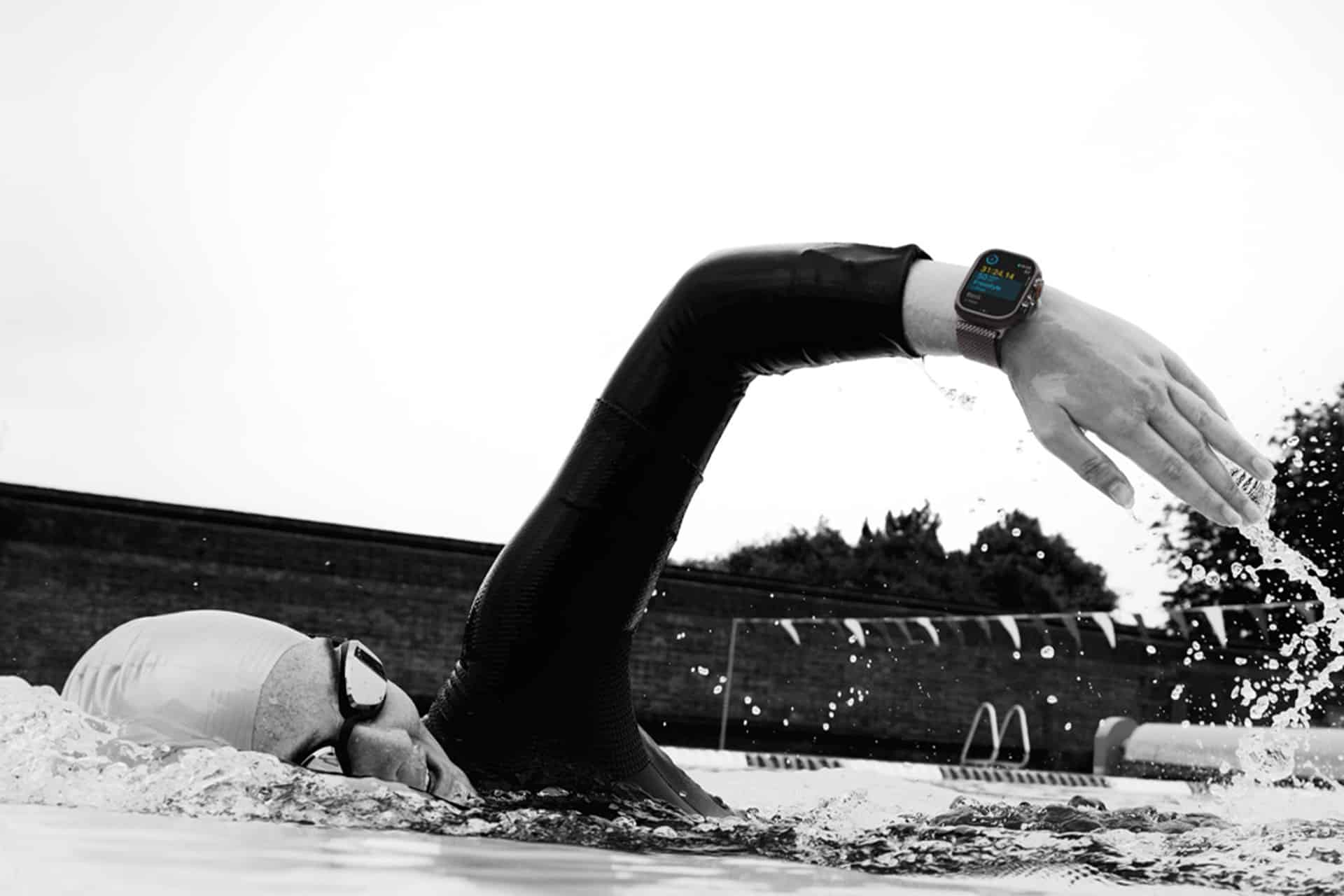 A swimmer in a pool performs a freestyle stroke, wearing goggles, a swim cap, and an Apple Watch Ultra displaying workout data on their wrist. The background features a fence, trees, and pool lane dividers.