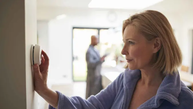 Mature Woman Turning Control Dial On Digital Central Heating Thermostat At Home.