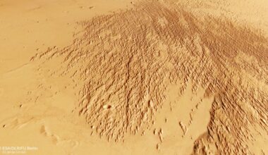 A section of Mars&rsquo;s surface showing a broad area covered with dense, elongated ridges and grooves that fan out diagonally across the image. The terrain is rough and textured in this region, contrasting with the smoother, lighter-coloured surrounding plains. The overall colour is a mix of tan and reddish-brown tones.