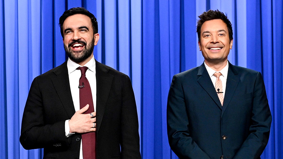 Zohran Mamdani stands beside Jimmy Fallon on a television stage during a late-night show monologue.