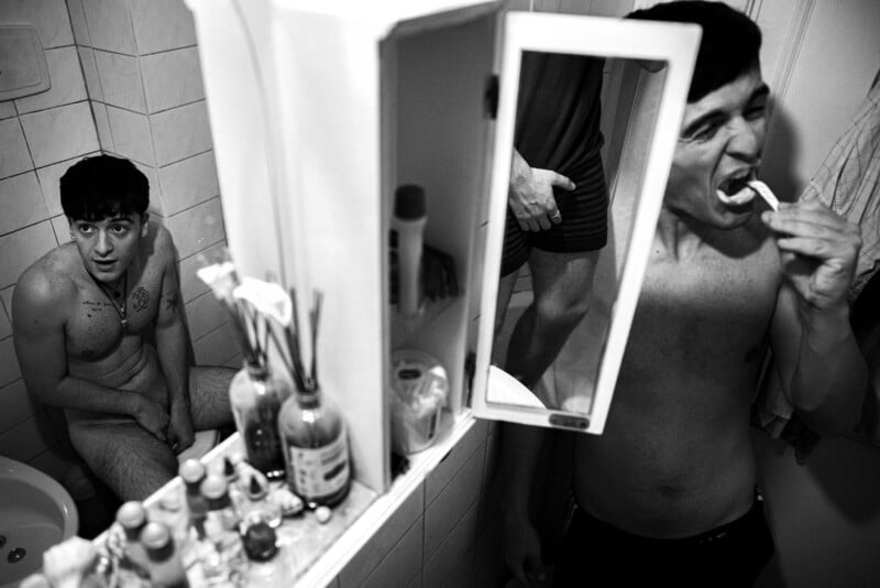 Two men in a small bathroom: one is sitting nude on the toilet, looking up, while the other brushes his teeth in front of the mirror. Toiletries clutter the sink and shelf, and the scene is in black and white.