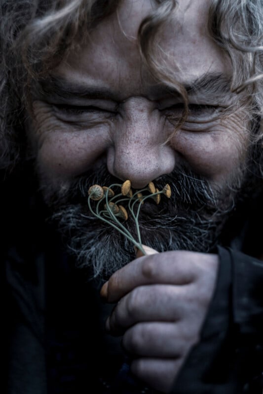 A bearded man with messy curly hair smiles joyfully, holding small yellow wildflowers close to his nose as if smelling them. His face is weathered and expressive, with deep lines and a playful expression.