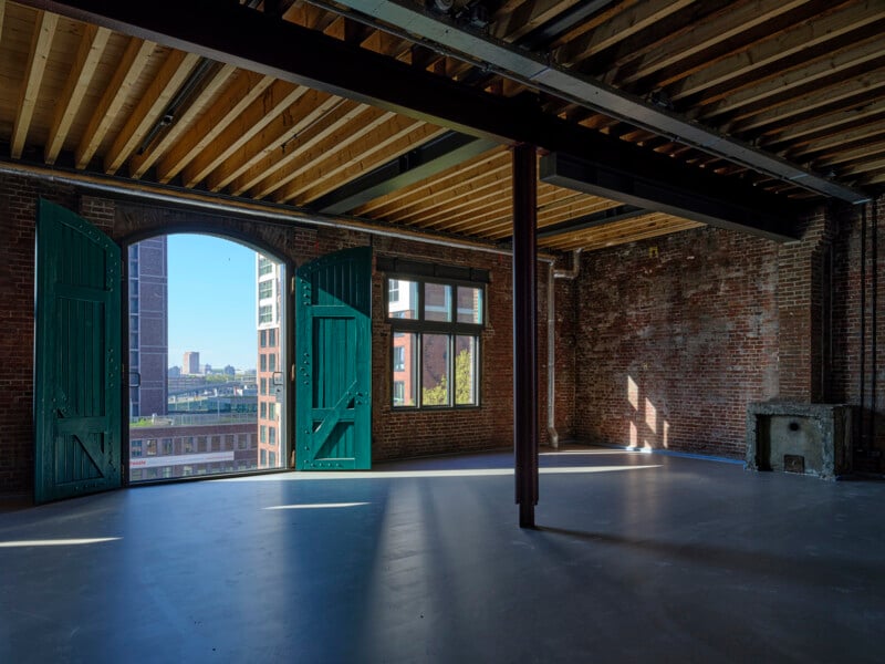 Sunlit industrial loft with exposed brick walls, wooden beam ceiling, open green double doors, large windows, and polished concrete floor. Cityscape and blue sky visible through the doors and windows.