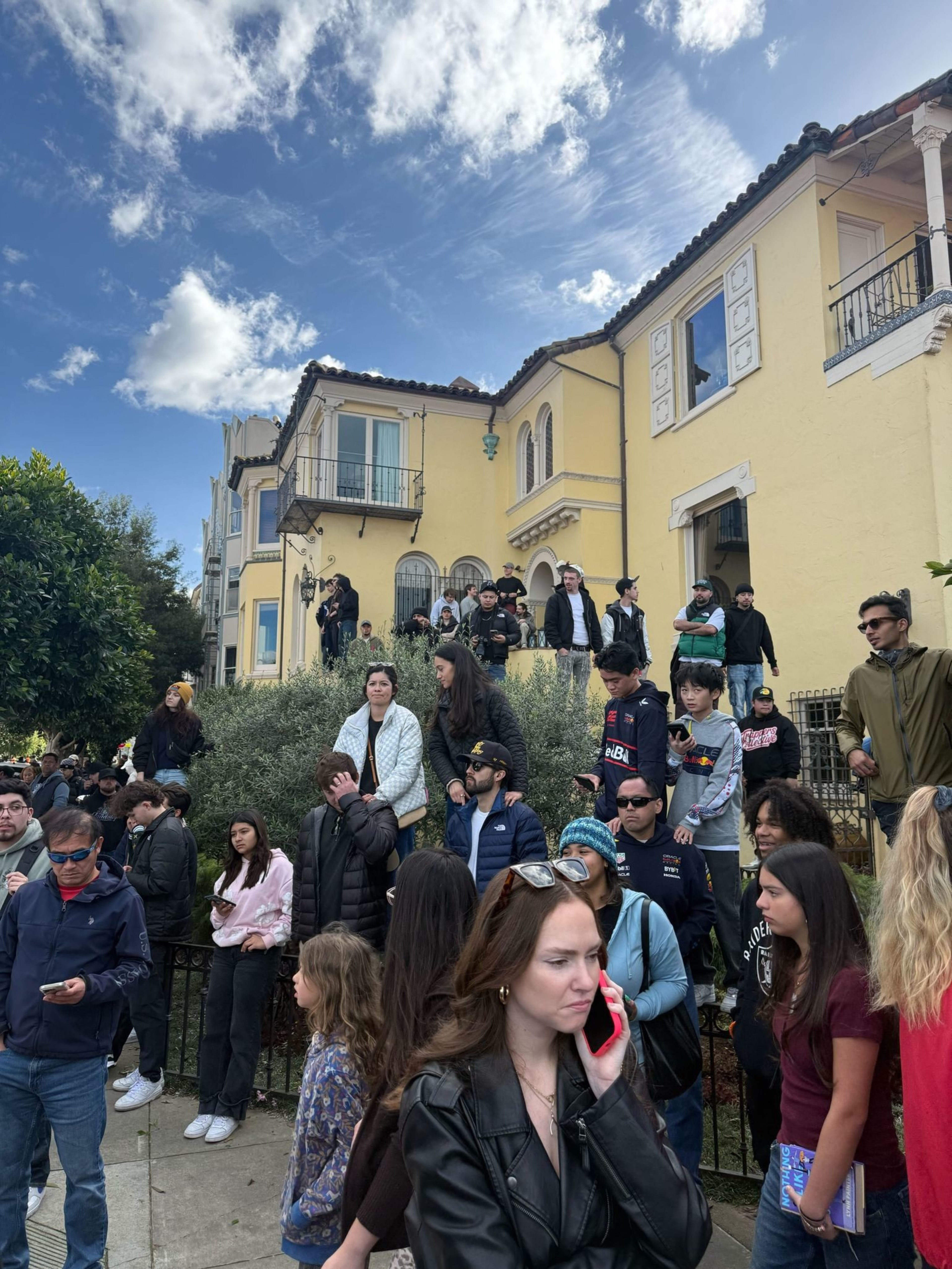 A crowd of people stands and sits outside a large yellow house under a partly cloudy blue sky, with some talking and others using phones.