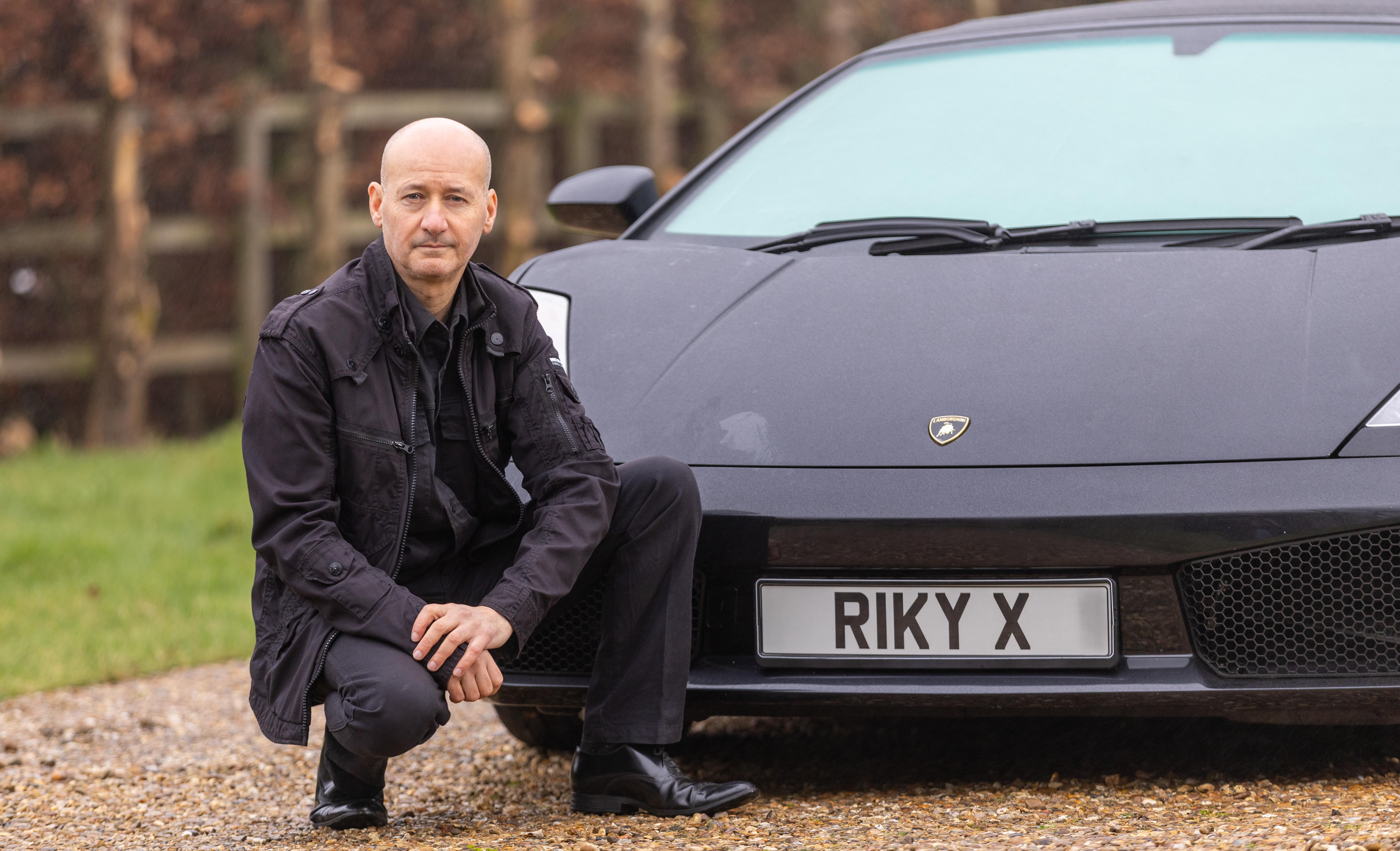 Stuntman Riky, 58, in a black jacket and pants, crouches next to a black Lamborghini with a personalized license plate that reads "RIKY X."