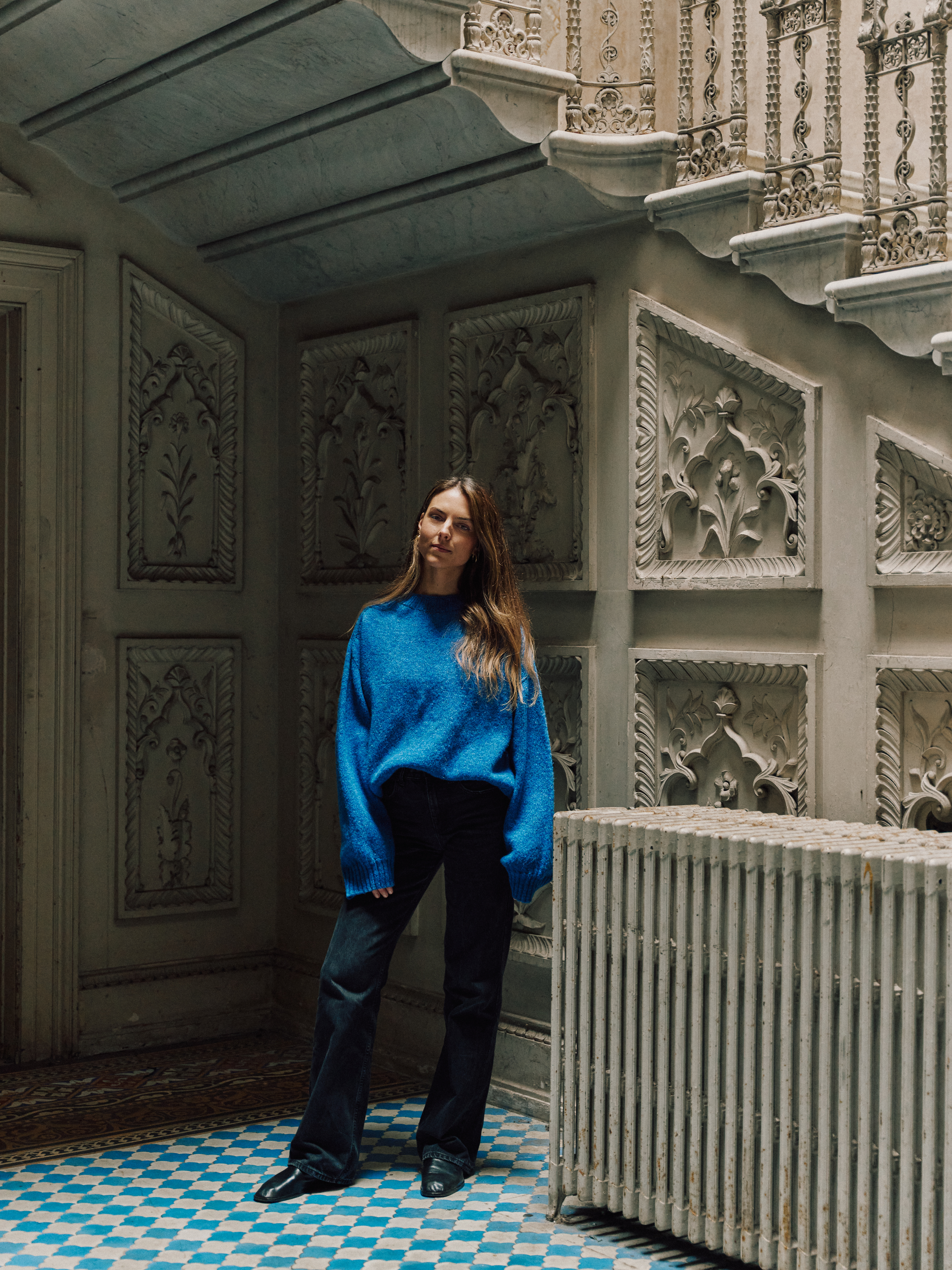 Phoebe McDowell in a blue sweater and black jeans standing in an ornate room with a patterned floor.