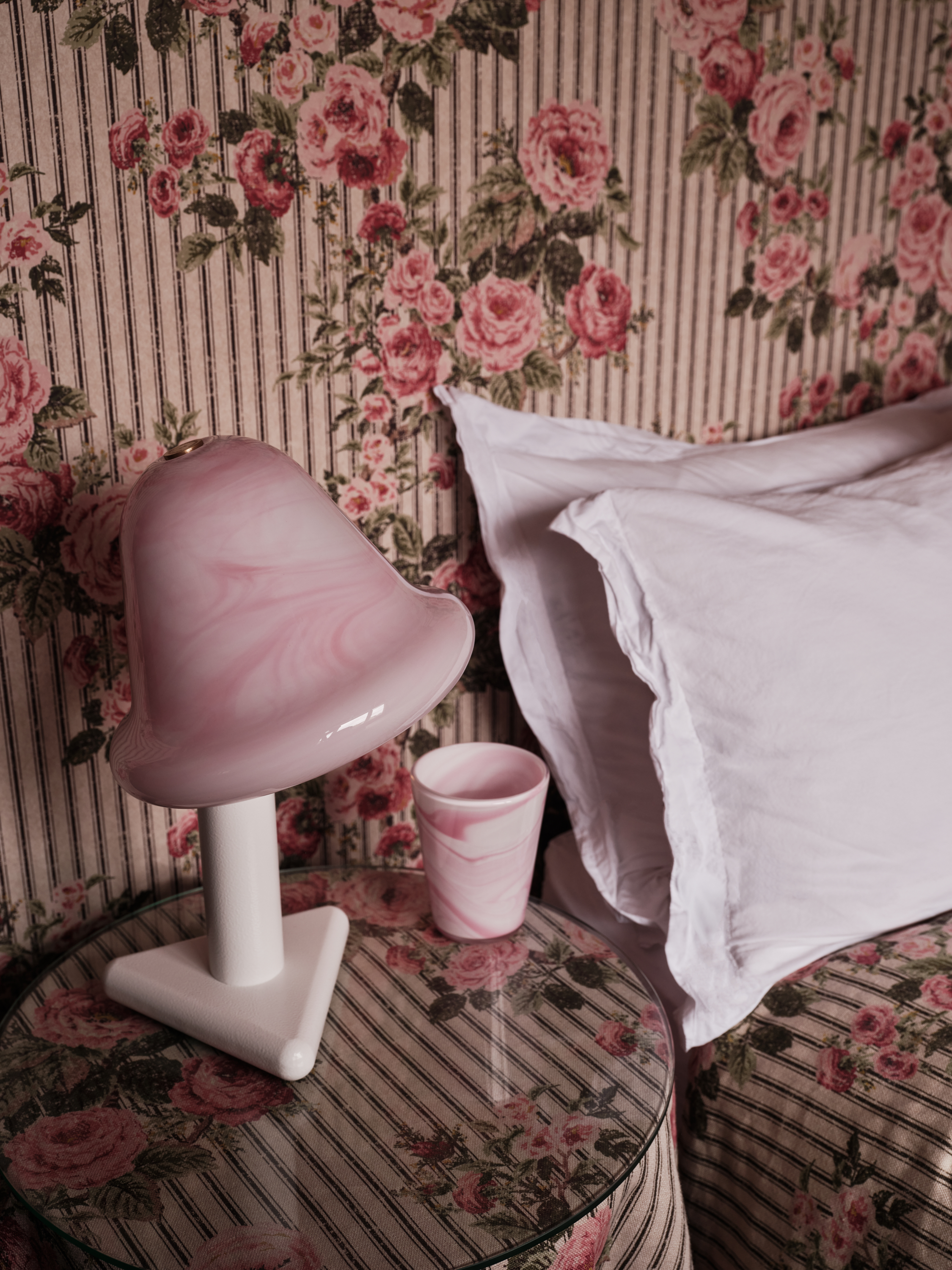 Pink swirled glass lamp and cup on a floral and striped patterned side table next to a bed.