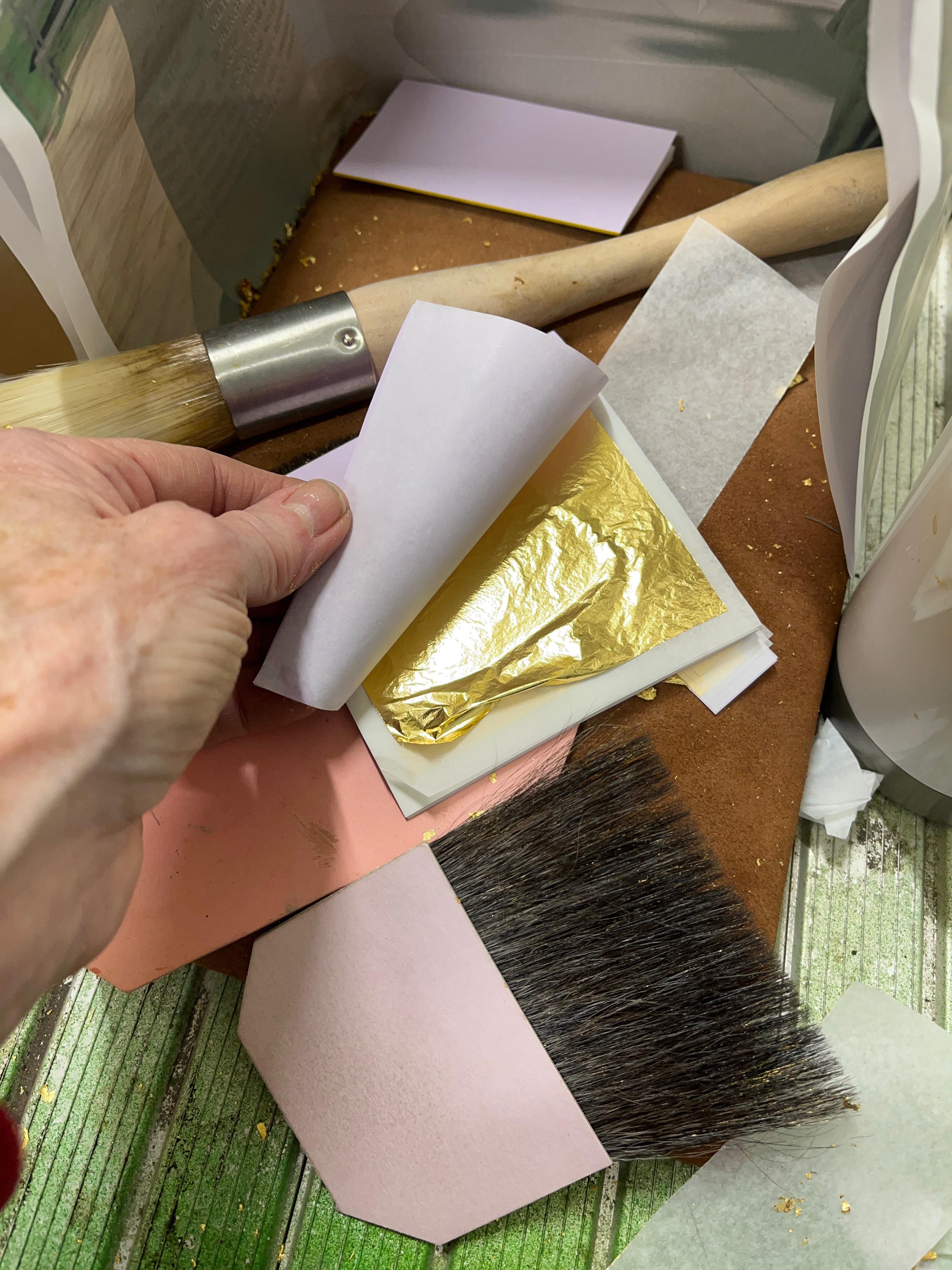 A person's hand opening a packet of gold leaf, surrounded by gilding tools and flakes of gold.