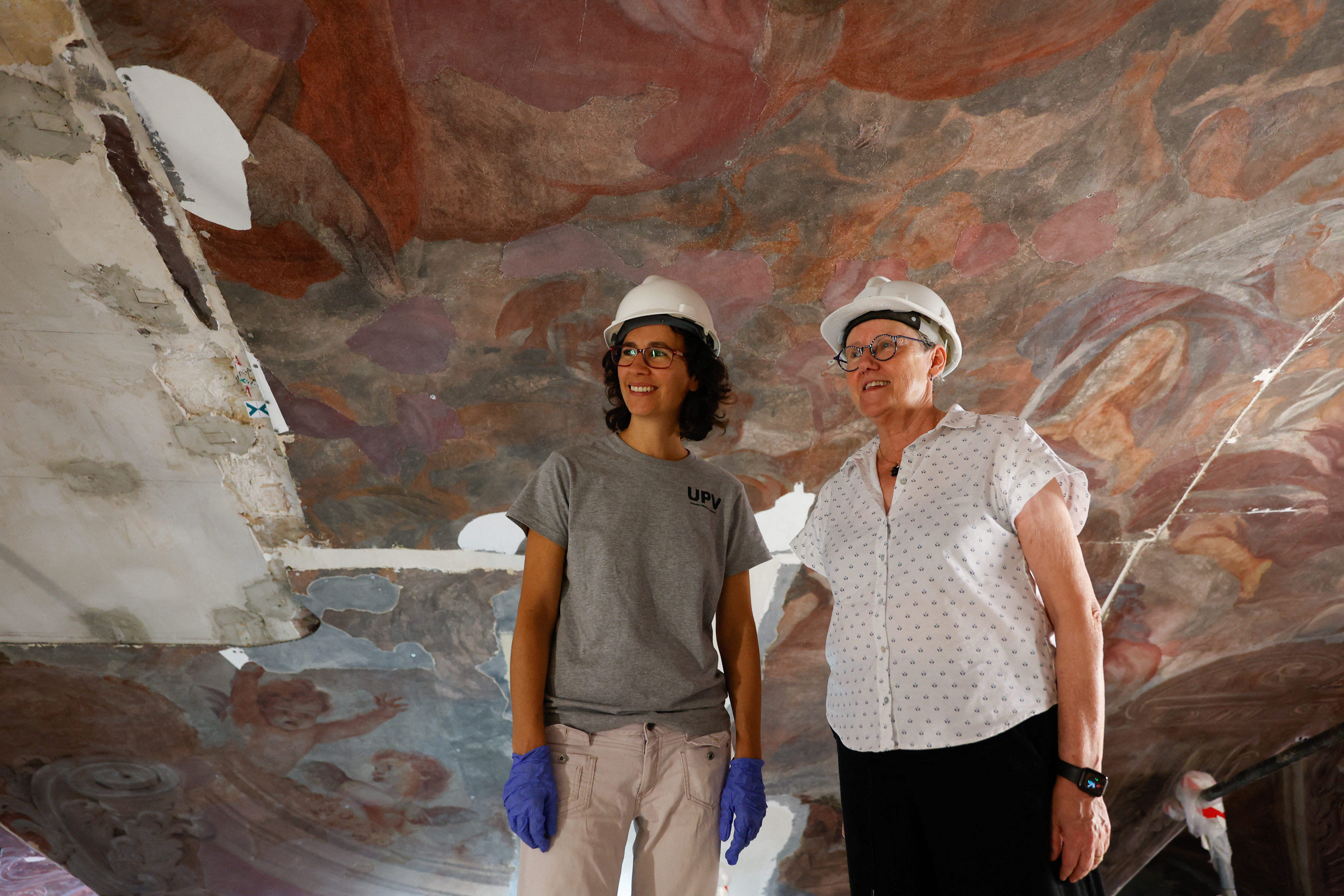 Two women in hard hats stand beneath a painted church ceiling in the process of restoration.